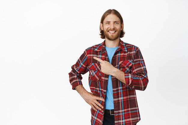 Retrato de hombre caucásico guapo con cabello rubio y barba, apuntando con el dedo en la esquina superior izquierda con una sonrisa feliz y orgullosa, expresión de cara entusiasta y segura en blanco