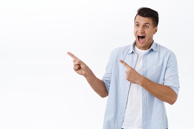 Retrato de un hombre caucásico guapo feliz emocionado reacciona alegre ante una oferta increíble señalando con el dedo a la izquierda y dejando caer la mandíbula al ver un nuevo artículo impresionante en stock de fondo blanco