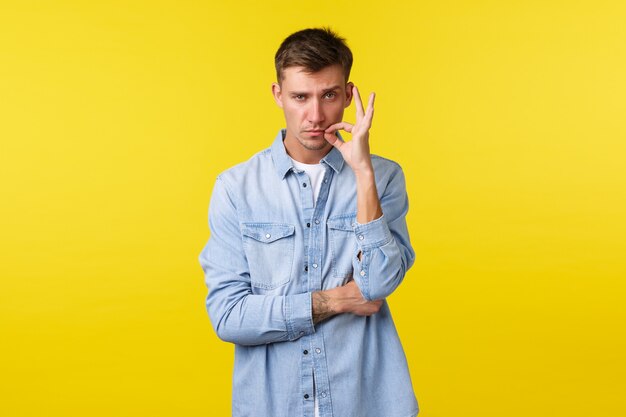 Retrato de hombre caucásico rubio de aspecto serio cerrando la boca con la cerradura, jurar mantener el secreto y permanecer en silencio, sellar los labios con promesa, de pie, decidido sobre fondo amarillo