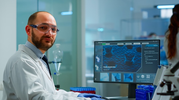 Retrato de hombre científico mirando a cámara sentado en laboratorio equipado moderno. Médico científico que examina la evolución del virus mediante mecanografía de alta tecnología en herramientas de química informática para la investigación científica