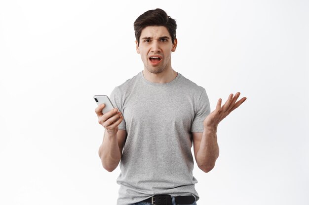 Retrato de hombre confundido con teléfono inteligente, sosteniendo el teléfono y quejándose de algo extraño, de pie desconcertado por el contenido en línea, fondo blanco.