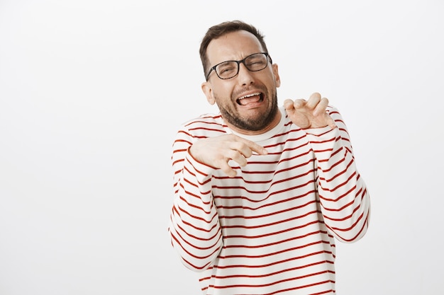 Retrato de hombre divertido y emotivo con gafas haciendo muecas e imitando patas de dinosaurio con las manos sobre el pecho