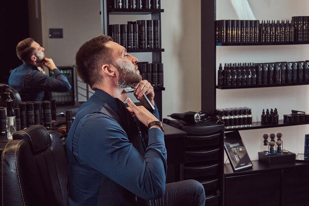 Retrato de un hombre elegante que se afeita mientras está sentado en una silla de peluquero en un salón de peluquería.