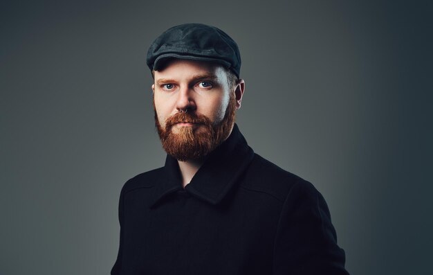 Retrato de hombre elegante Redbeard vestido con una gorra plana de tweed y una chaqueta negra sobre fondo gris.