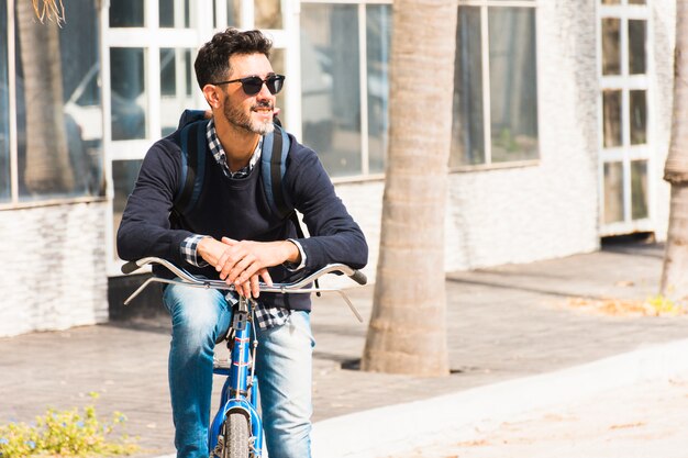 Retrato del hombre elegante sonriente con su mochila que se sienta en su bicicleta que mira lejos