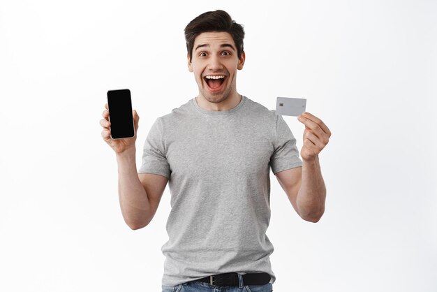 Retrato de un hombre emocionado que muestra la pantalla vacía del teléfono y la tarjeta de crédito demuestra la tienda en línea y hace un pedido de pie sobre fondo blanco