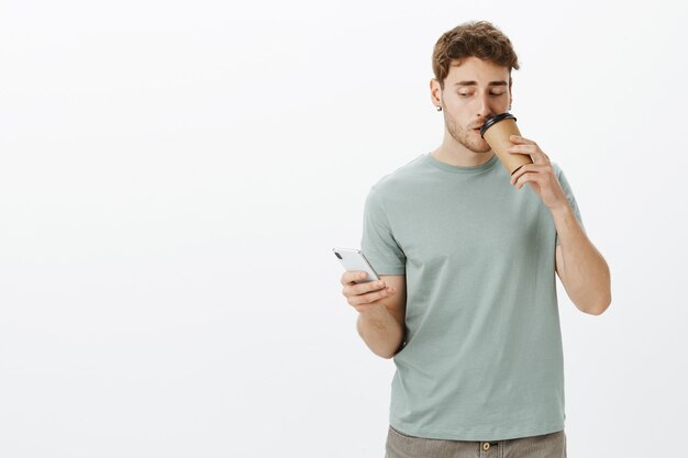 Retrato de hombre empresario guapo seguro en aretes, sosteniendo un teléfono inteligente y bebiendo una taza de café mientras responde el mensaje y mira la pantalla