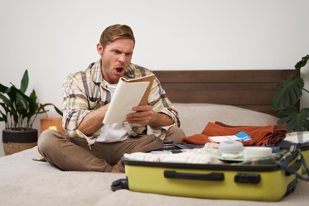 Foto gratuita retrato de un hombre enojado empacando una maleta mirando frustrado al cuaderno con una lista de comprobación de artículos para