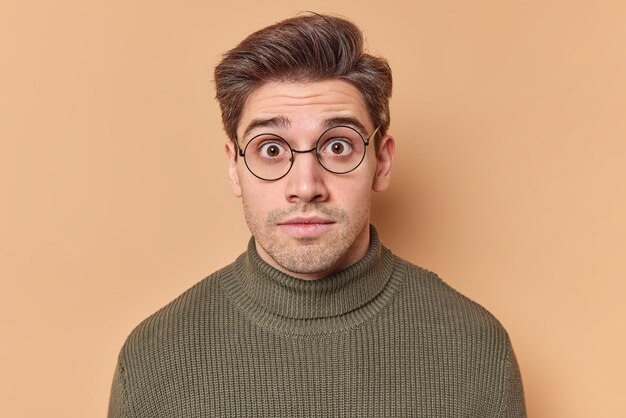 Retrato de un hombre europeo brunet sorprendido que mira fijamente a la cámara, mantiene los ojos bien abiertos, lleva gafas redondas y un suéter ve algo impresionante aislado sobre fondo beige de estudio