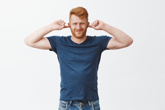 Retrato de hombre europeo fuerte atractivo disgustado con cabello pelirrojo, cubriéndose las orejas, frunciendo el ceño y frunciendo los labios por la incomodidad, molesto con el sonido fuerte