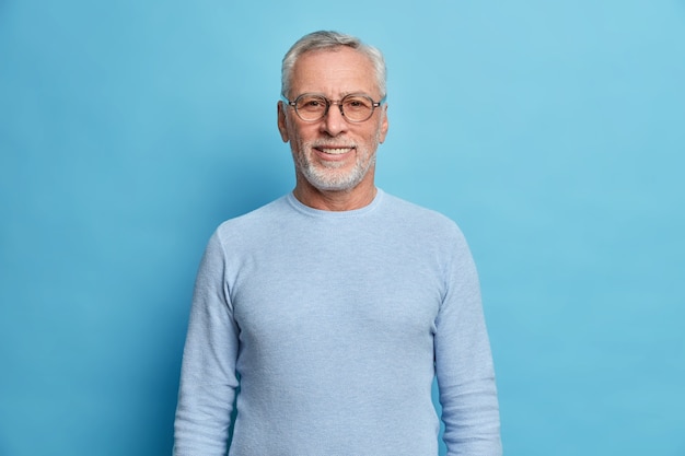 Retrato de hombre europeo guapo con barba con cabello gris y barba sonríe agradablemente mira directamente al frente estando de buen humor tiene día de suerte usa anteojos y suéter aislado sobre pared azul