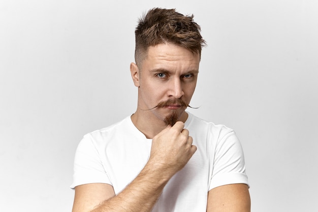 Retrato de hombre europeo joven serio sospechoso en camiseta blanca mirando a la cámara con sospecha y desconfianza, sosteniendo la mano en su barba. Chico barbudo pensativo con elegante bigote posando en interiores