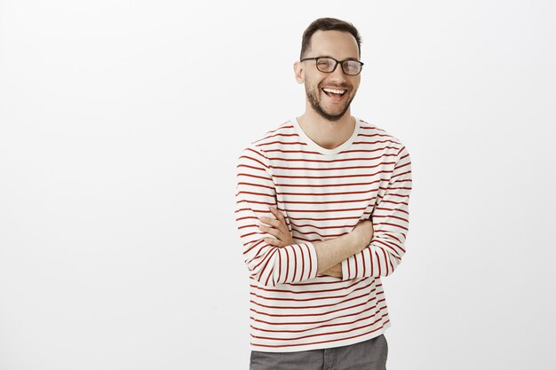 Retrato de hombre europeo tímido y guapo con barba con gafas, cruzando las manos sobre el pecho y riendo en voz alta, sintiéndose genial mientras habla casualmente con un nuevo grupo de amigos sobre una pared gris