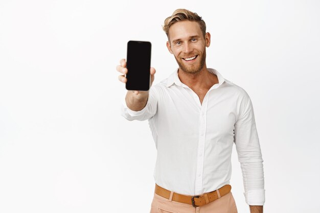Retrato de un hombre exitoso sonriente que muestra un teléfono celular con una pantalla negra vacía que muestra un producto o una tienda en línea en el fondo blanco del teléfono móvil