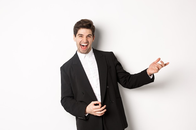 Retrato de hombre feliz bailando en la fiesta, tocando la guitarra invisible y riendo, de pie en traje negro sobre fondo blanco.