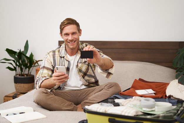 Foto gratuita retrato de un hombre feliz comprando boletos con devolución de tarjeta de crédito sentado en la cama y empacando ropa