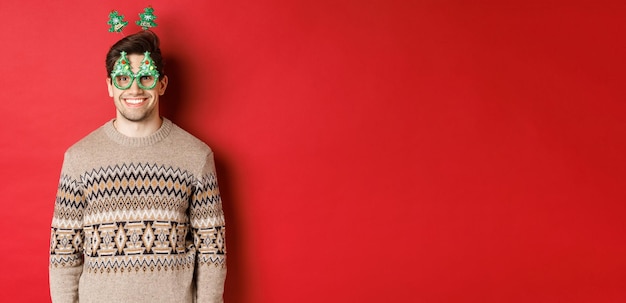 Retrato de hombre feliz y lindo en suéter de invierno y gafas de fiesta celebrando año nuevo o Navidad sonriendo mientras está de pie sobre fondo rojo