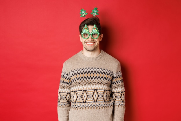 Retrato de hombre feliz y lindo en suéter de invierno y gafas de fiesta, celebrando el año nuevo o la Navidad, sonriendo mientras está de pie sobre fondo rojo.