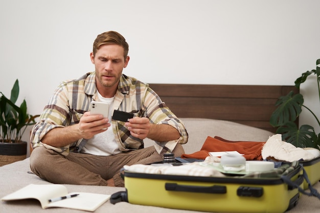 Foto gratuita retrato de un hombre feliz en una maleta transfiriendo dinero comprando boletos en línea sentado en la cama con