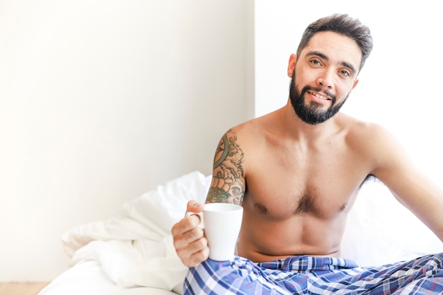 Retrato de un hombre feliz sentado en la cama sosteniendo una taza de café