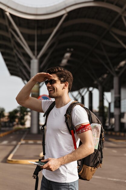 Retrato de hombre fresco en camiseta blanca mirando a la distancia y sonriente Chico morena con gafas de sol sonríe y tiene pasaporte cerca del aeropuerto