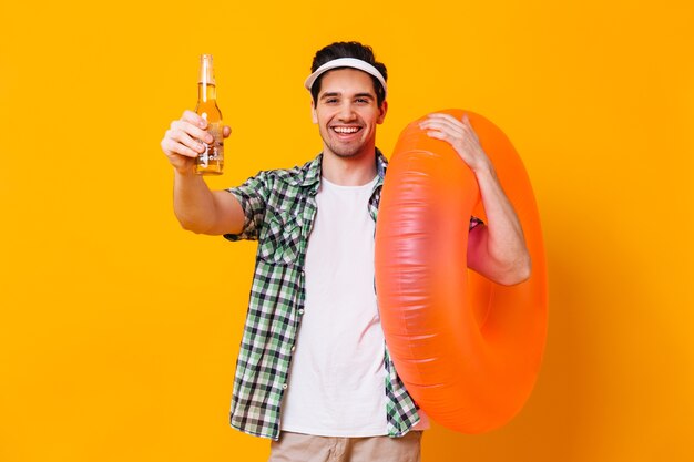 Retrato de hombre con gorra, camisa y camiseta sosteniendo una botella de cerveza y un círculo inflable en un espacio aislado.