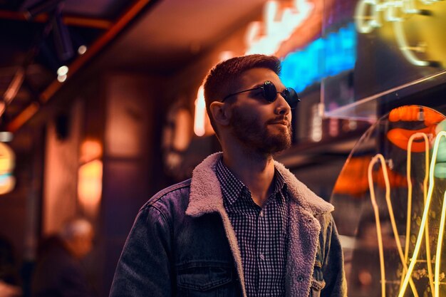 Retrato de un hombre guapo con un abrigo de mezclilla y gafas de sol parados en la noche en la calle. Letreros iluminados, neón, luces.