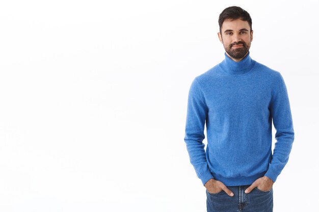 Retrato de hombre guapo adulto con barba, tomarse de las manos en los bolsillos de los pantalones vaqueros mientras sonríe con expresión amistosa y despreocupada, de pie en la pared blanca pose normal