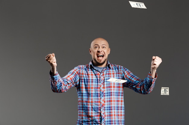 Foto gratuita retrato de hombre guapo afortunado en camisa a cuadros agitando las manos gritando sobre la pared gris con dinero cayendo desde arriba