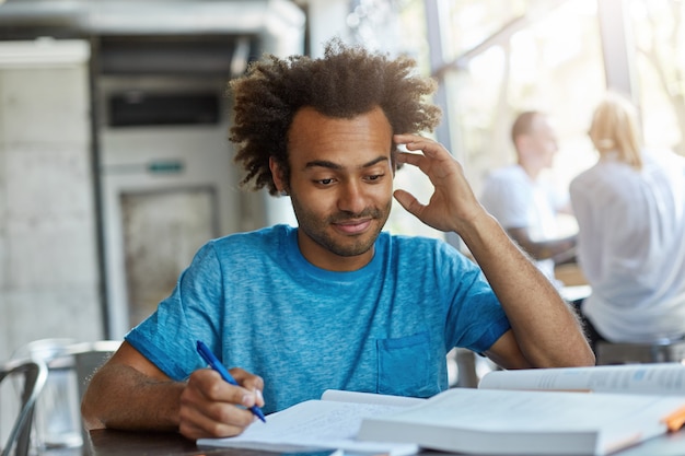 Retrato de hombre guapo afroamericano con cabello tupido sentado en el escritorio en la cantina de la universidad escribiendo notas rascándose la cabeza sin saber algo que prepara una investigación o proyecto científico