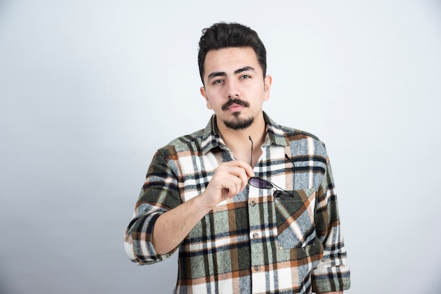 Retrato de hombre guapo con barba y gafas posando y de pie sobre la pared blanca.