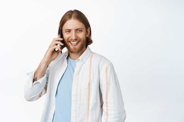 Retrato de hombre guapo con barba hablando por teléfono móvil y sonriendo, riendo casualmente en blanco
