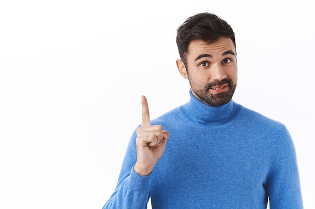 Retrato de hombre guapo con barba inteligente agrega una solución, tiene una idea o sugerencia, levanta un dedo para decir una recomendación, da un consejo mientras está parado sobre una pared blanca