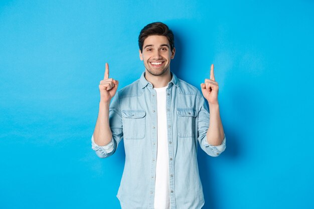 Retrato de hombre guapo con barba en traje casual, sonriendo feliz y apuntando con el dedo hacia arriba en el espacio de la copia, de pie sobre fondo azul.