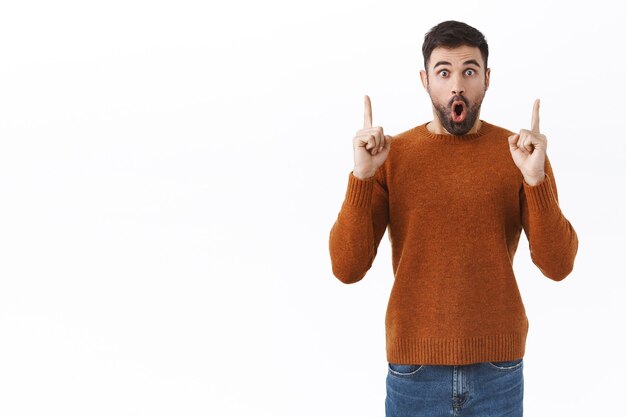 Retrato de hombre guapo barbudo sorprendido y sorprendido, jadeando sorprendido con incredulidad mientras señala con el dedo hacia abajo, mostrando nuevas e increíbles noticias frescas, de pie en la pared blanca