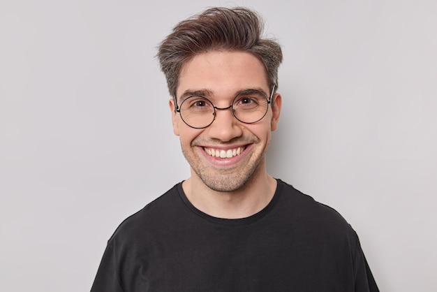 Retrato de hombre guapo brunet sonríe alegre viste camiseta negra básica y gafas redondas mira directamente a la cámara tiene una conversación feliz aislada sobre fondo blanco. Emociones felices.