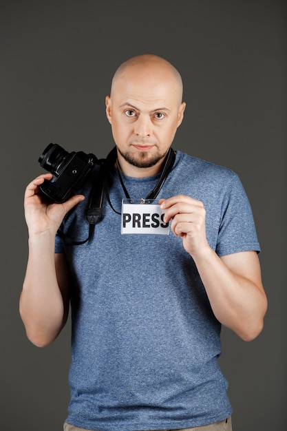 Retrato de hombre guapo en camisa gris con cámaras y placa de prensa posando sobre pared oscura