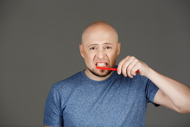 Retrato de hombre guapo divertido en camisa gris cepillarse los dientes sobre la pared oscura