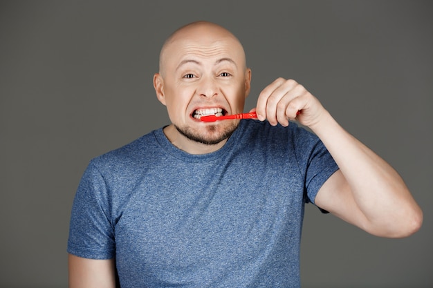 Retrato de hombre guapo divertido en camisa gris cepillarse los dientes sobre la pared oscura