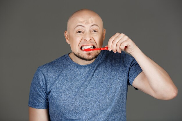 Retrato de hombre guapo divertido en camisa gris cepillarse los dientes sobre la pared oscura