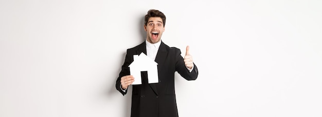 Foto gratuita retrato de un hombre guapo emocionado que busca una casa que muestre una casa y un pulgar recomendando bienes raíces