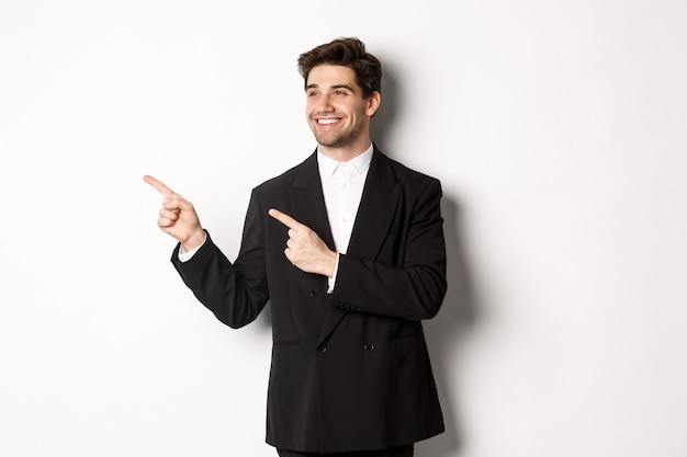 Retrato de hombre guapo exitoso en traje, apuntando y mirando a la izquierda con sonrisa complacida, mostrando banner promocional, de pie sobre fondo blanco.