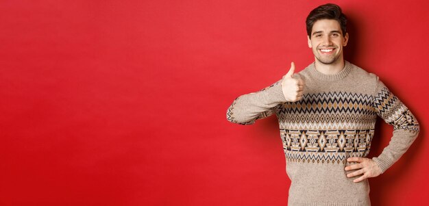 Retrato de hombre guapo feliz en suéter de navidad sonriendo y luciendo satisfecho mostrando pulgares arriba en aprobación como vacaciones de año nuevo de pie sobre fondo rojo