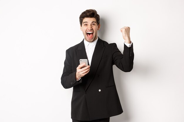 Retrato de hombre guapo feliz en traje, regocijo, lograr el objetivo en la aplicación móvil, levantando el puño y gritando que sí, sosteniendo el teléfono inteligente, de pie contra el fondo blanco