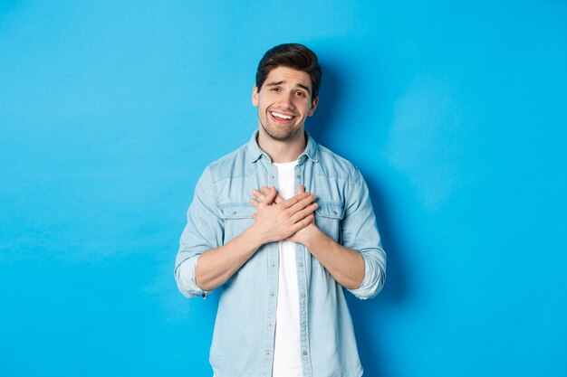 Retrato de hombre guapo halagado agradeciéndole, tomados de la mano en el corazón y sonriendo, mirando agradecido, de pie sobre fondo azul.