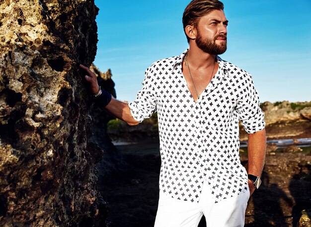 Retrato de hombre guapo modelo vistiendo ropa blanca posando cerca de rocas en la playa en el cielo azul