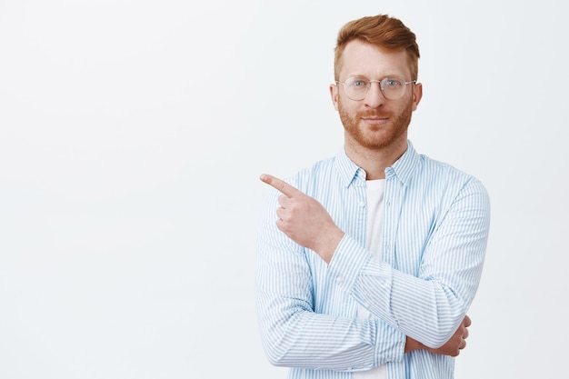 Retrato de hombre guapo pelirrojo con barba en gafas y camisa apuntando a la esquina superior izquierda y sonriendo