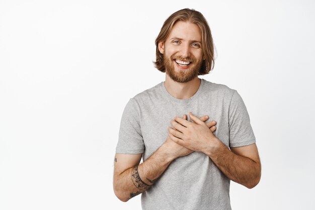 Retrato de hombre guapo rubio con barba, sonriendo y tomados de la mano en el corazón, agradeciéndole, siendo halagado, agradecido, expresando gratitud, de pie en camiseta gris sobre blanco