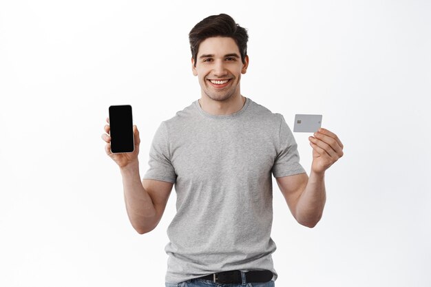 Retrato de hombre guapo satisfecho que muestra la pantalla vacía del teléfono inteligente y la tarjeta de crédito plástica, demuestra la aplicación del teléfono, el concepto de banca y finanzas