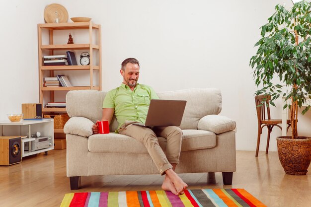 Retrato de hombre guapo sentado en un sofá o sofá y trabajando en la computadora portátil.